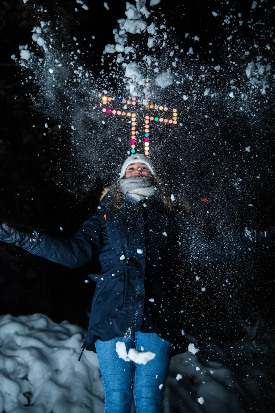Une personne lance de la neige dans la nuit, devant des lumières colorées.